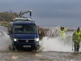 A van is driven through floodwater, splashing two men in hi-vis waterproofs walking nearby